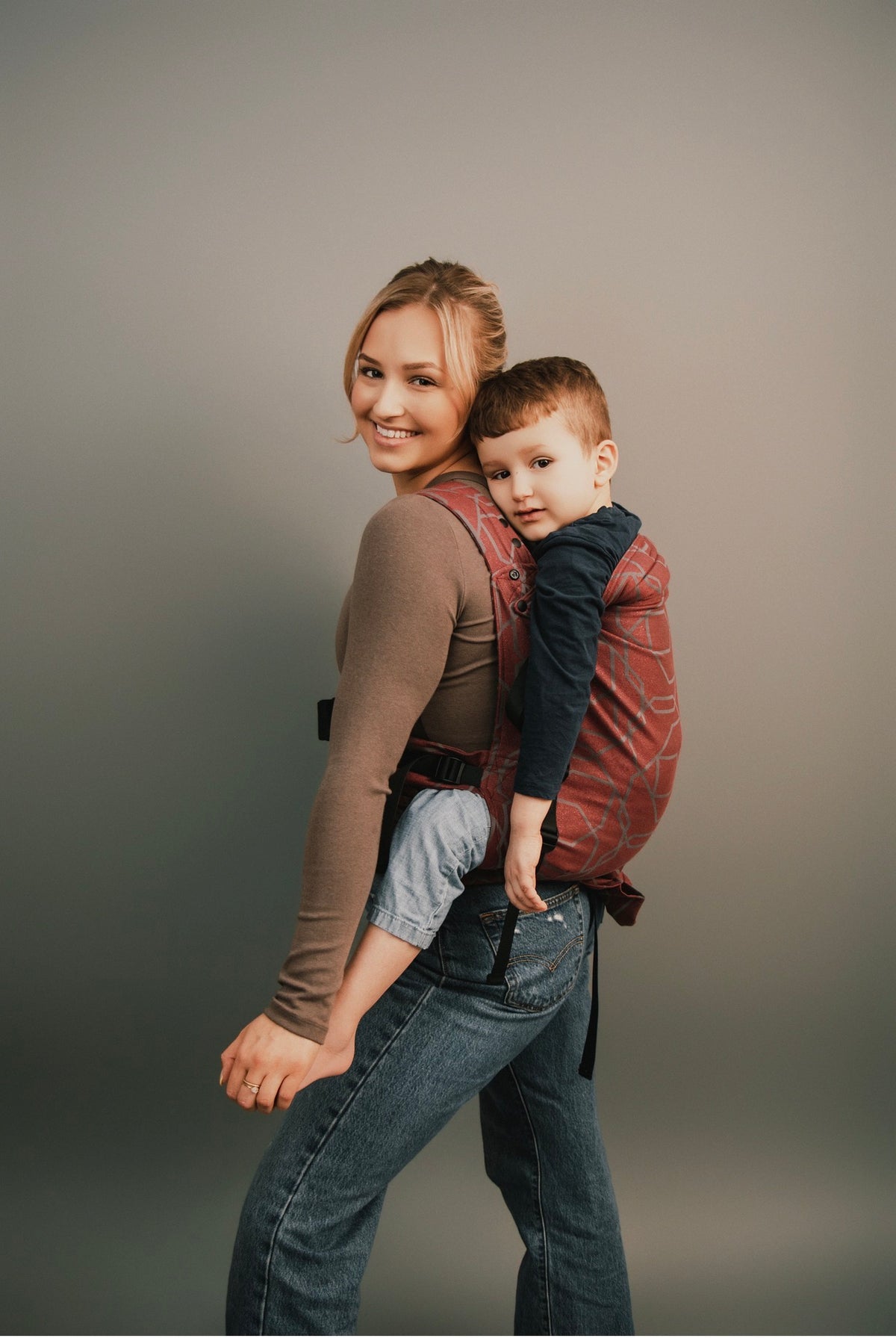 A woman wearing a brown long-sleeve top and blue jeans carries a young boy on her back in a Gogo against a plain gray studio background.