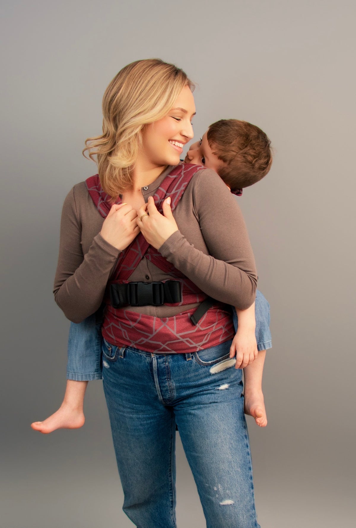 A woman wearing a Gogi carrier, carrying a child on her back, photographed against a solid gray studio background.