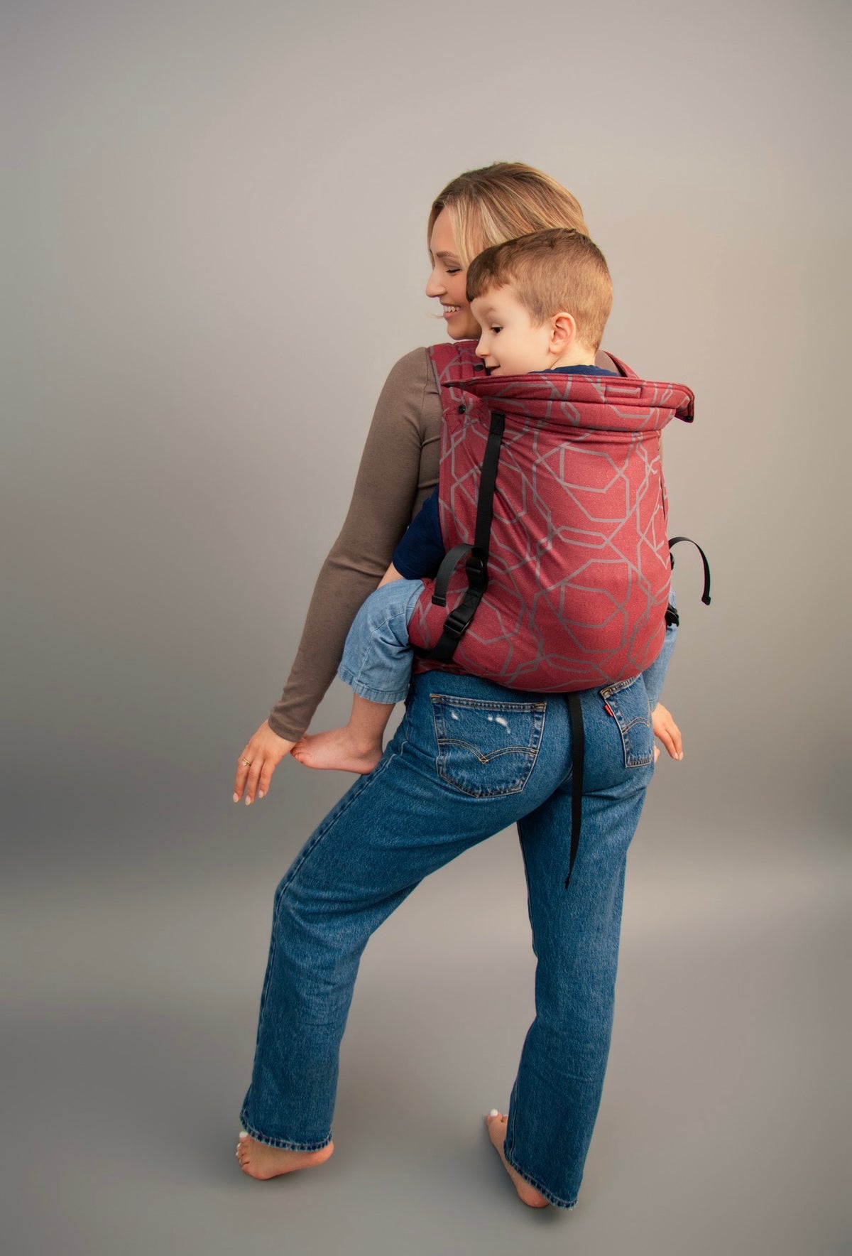 A woman wearing blue jeans and a brown long-sleeve top carries a young child in a Gogo carrier, photographed against a neutral gray studio background.