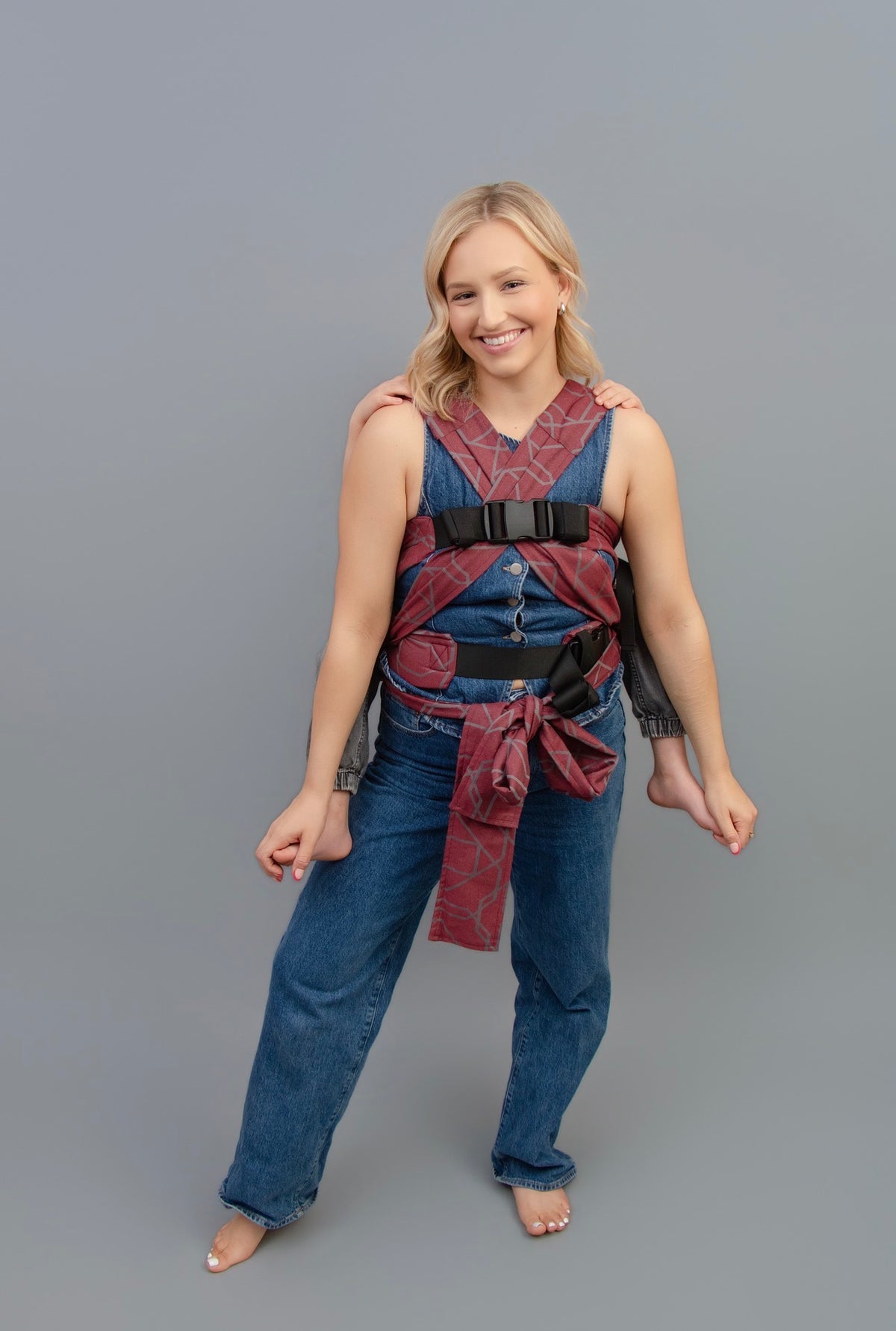 A woman with blonde hair is front facing, wearing a child on her back with a Gogo carrier, standing against a solid gray studio background.