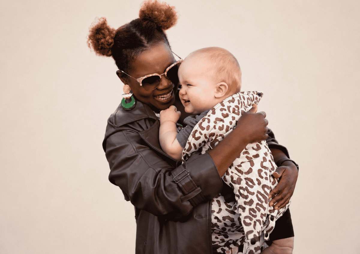 A woman in a dark brown leather jacket and sunglasses holds a baby in an Mbererko carrier against a solid light beige background.