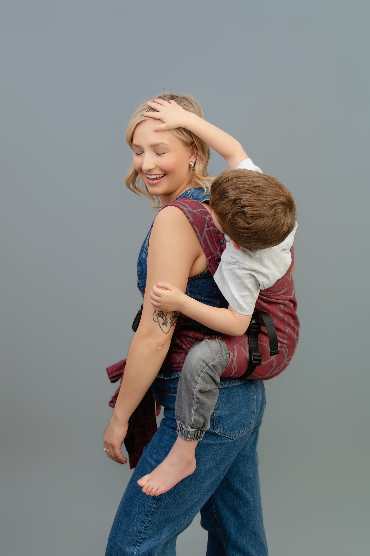 A woman wearing a Gogo carrier with a young child sitting on her back, photographed against a solid gray studio background.